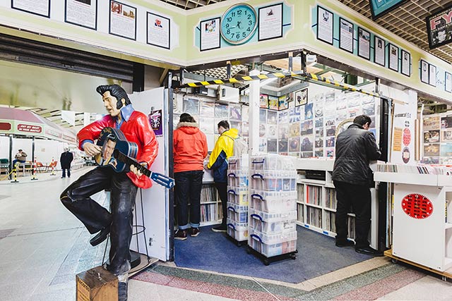 Indoor market stall containing vinyl records. The image also contains a statue of Elvis in a red jacket black trousers holding a black guitar with an image of Elvis on the front  