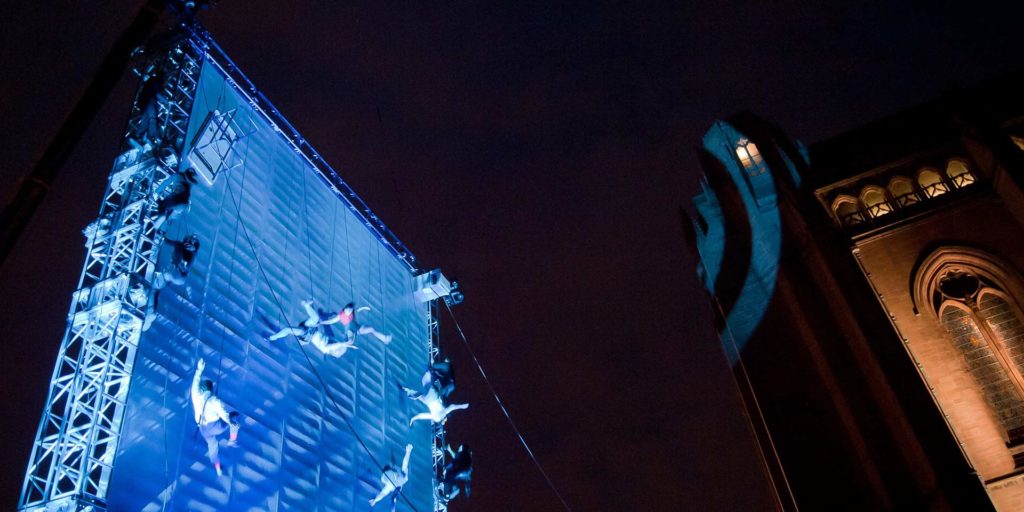 Wired acrobats appearing to fall from blue structure against black night sky