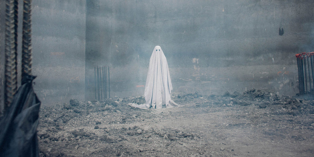 A person wearing a white sheet dressed as a ghost, standing against a misty background. The eerie atmosphere is enhanced by the fog, creating a spooky and mysterious effect.