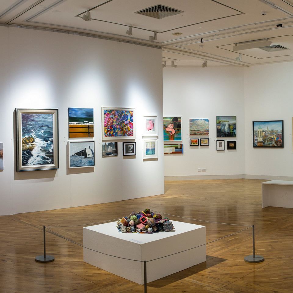 Large room painted white with vaious sized modern painting long the central band. a low platform in the center with pile of multi colored object in the center surrounded by a low metal rope barrier.