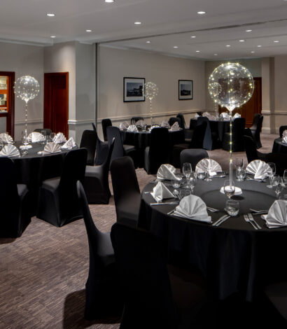 Function room with 4 dining tables set for 32 people. Decorated in Black and silver with fan serviette and a single transparent balloon floating above each table. The room has a Mirrored wall and private bar