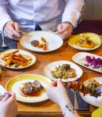 Wooden table with various Tapas on plates hands holding knives and forks