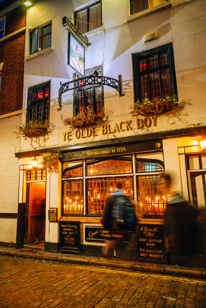 in Gold Letters Ye Olde Back Boy, White painted building with black window frames. Flowers in window boxes in upper floors, blurred people in the foreground.
