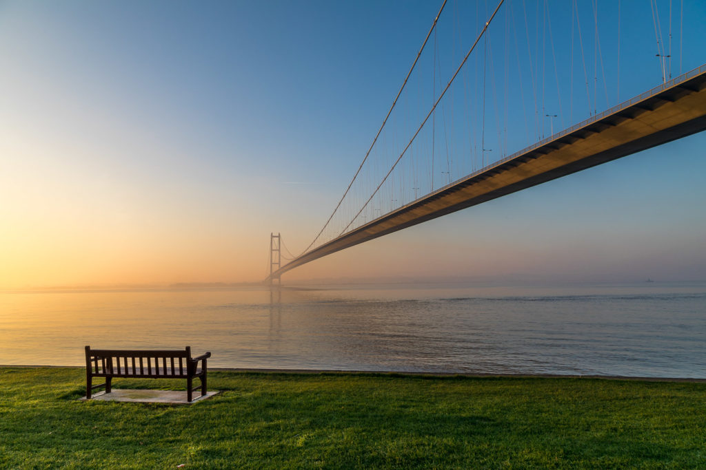 Single span suspension bridge over large body of water. Sunrise on a misty day. There is also a bench in the foreground on a grassy bank