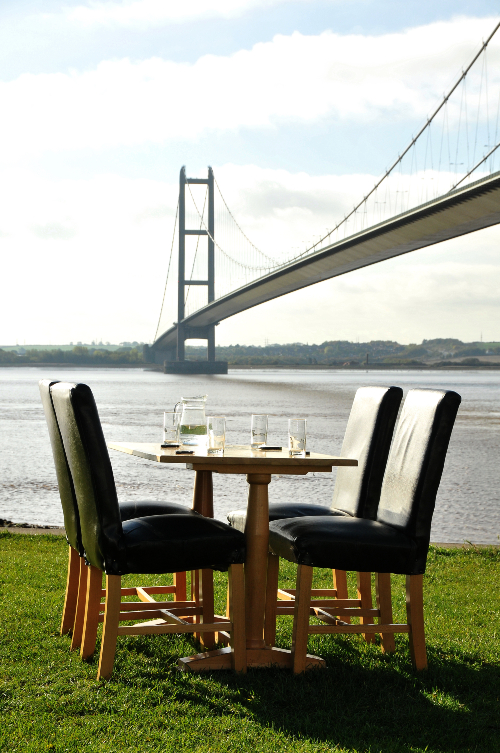 Table and 4 chairs set for dinner in a grassed area with the Humber Bridge in the background