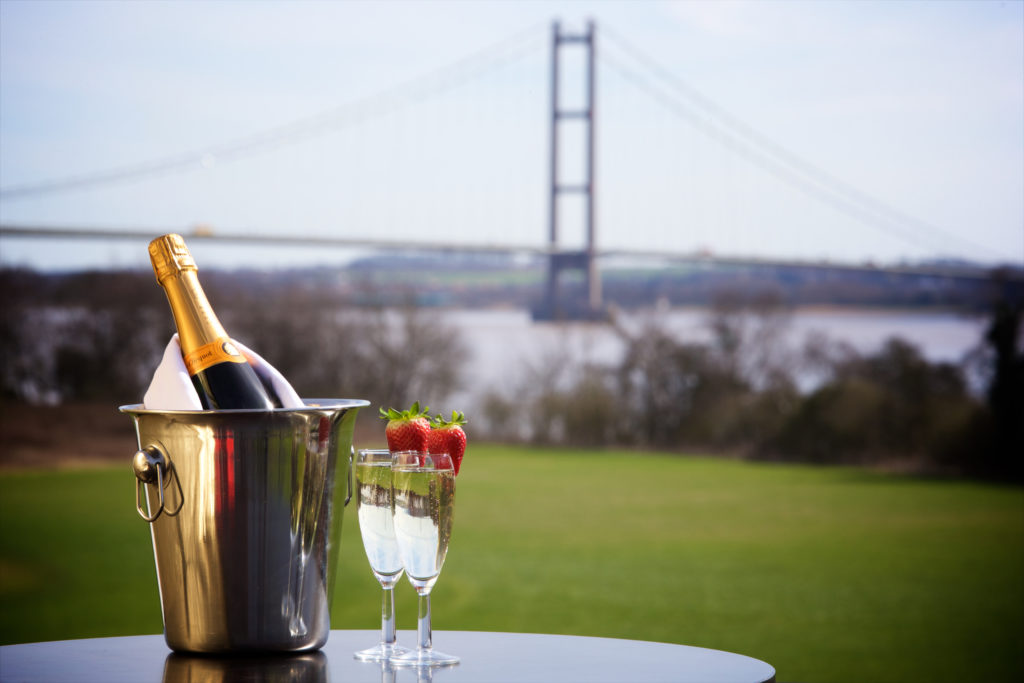 Bottle of Champagne in silver bucket alongside 1 Champagne flutes topped with strawberries. Lawn and Humber bridge in the background