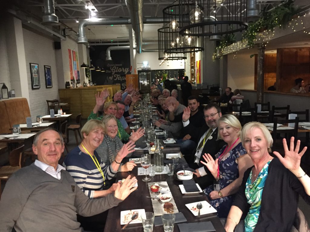 Group of people at a long table enjoying a meal smiling and waving 