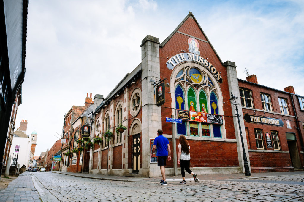 Red brick building with steep pitched roof. The works The Mission over large ached stain glass window. Cobbled street and 2 people in the foreground