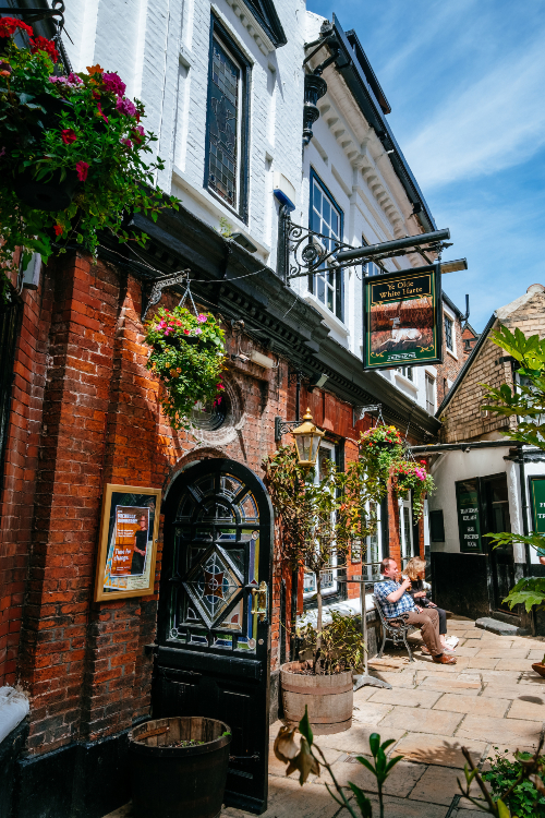 Red brick courtyard with lots of summer hanging baskets and flowers. 2 people are sitting on a bench.