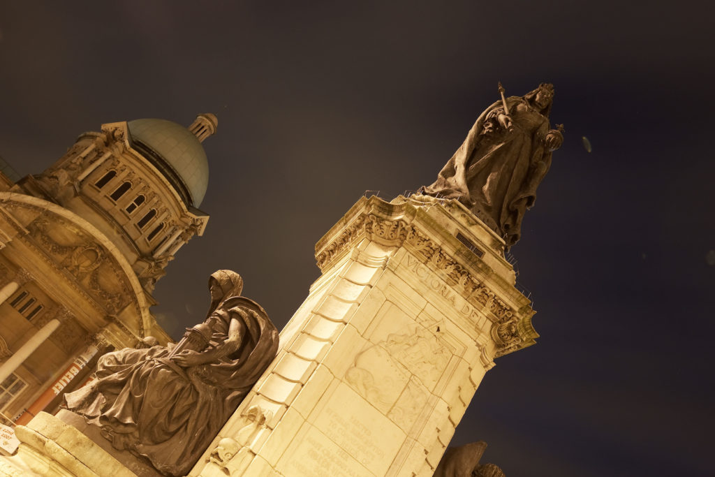 Alt text: "A dramatic nighttime photograph of the Queen Victoria statue in Queen Victoria Square, Hull. The statue is illuminated against the dark sky, with the grand facade of Hull City Hall glowing in the background, highlighting the city's historic