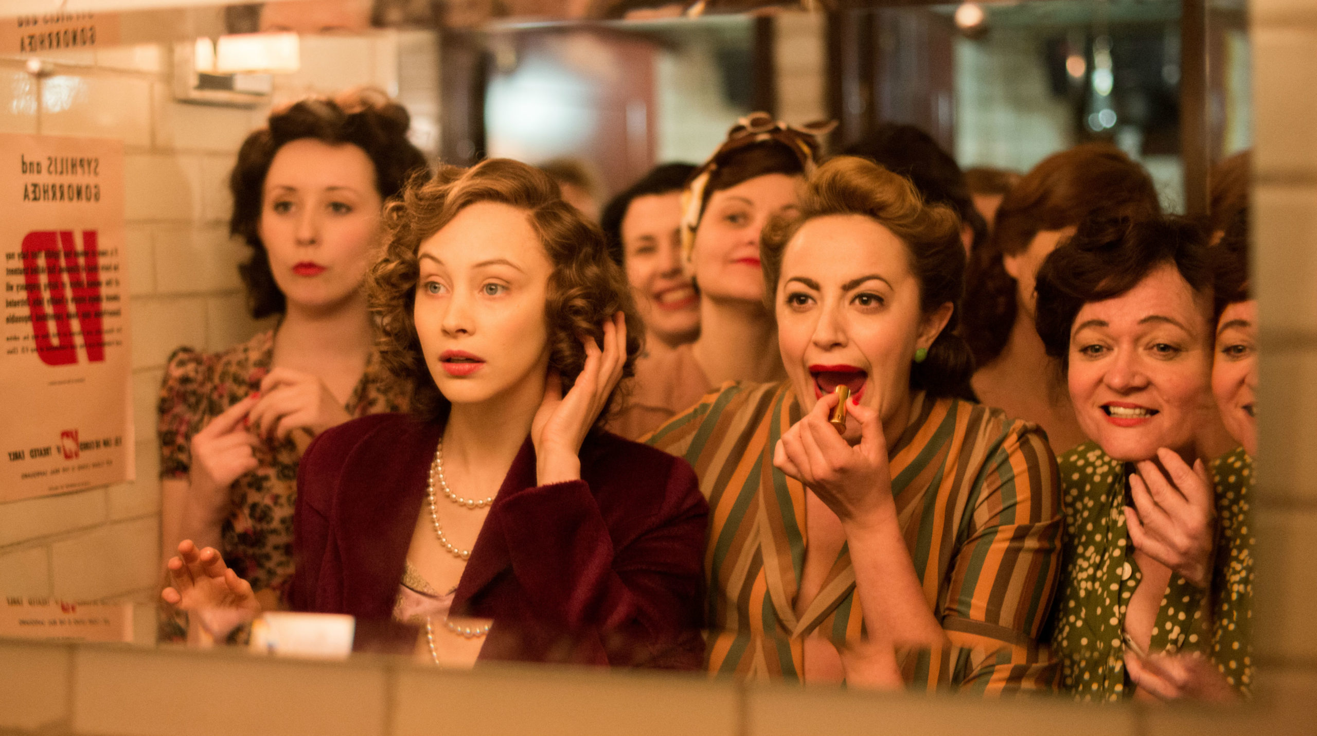 A group of women in 1950 attire looking in a mirror applying makeup and fixing their hair