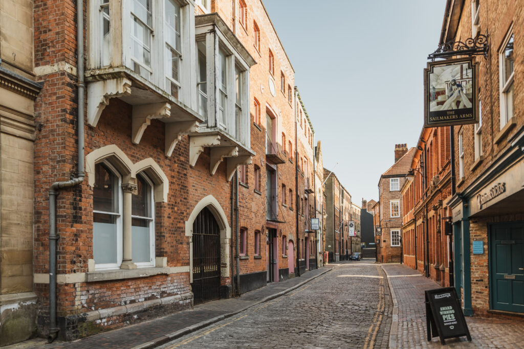 Hull's High Street, lined with old brick buildings, traditional shopfronts, and cobbled pavements. The street has a mix of heritage architecture and modern elements, reflecting the city's rich maritime and trading history.