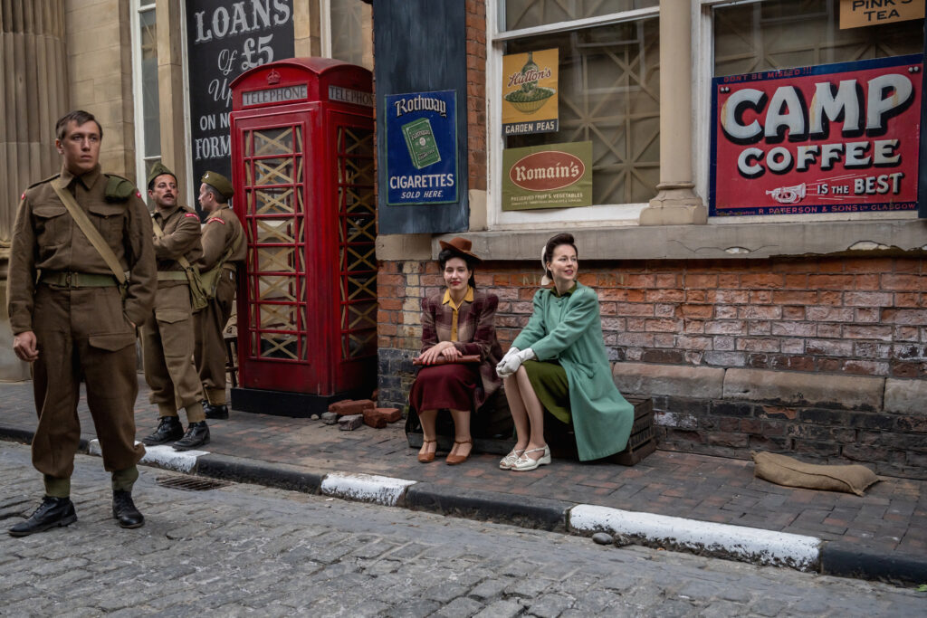 Filming of The Crown on Hull's High Street in October 2022. Two women in elegant period dresses stand near men in vintage army uniform. The historic street, lined with old brick buildings, is transformed with props and set pieces to reflect the era being depicted in the production