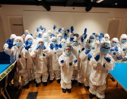 A group of children taking part in science activities, they are dressed in white hazmat suits with masks and gloves on.