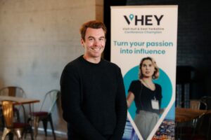 Ryan Shaw is stood in front of a blue banner that says "VHEY Conference Champions Turn Your Passion Into Influence". He has short red hair and is smiling and wearing a black top and black jacket.
