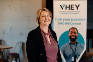 Samanth Dunion is stood in front of a blue banner that says "VHEY Conference Champions Turn Your Passion Into Influence". She has a blonde bob, is wearing a pink blouse and black jacket and is smiling.