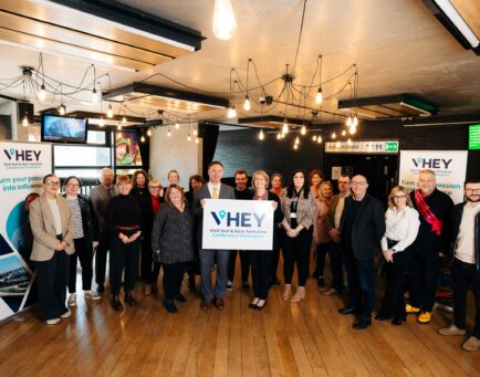 Conference Champions gathered in Hull Truck Theatre to celebrate launch. There is a mix of ages and genders. Everyone is smiling and in the centre a delegate is holding a white board with "VHEY Conference Champions" branded in blue.
