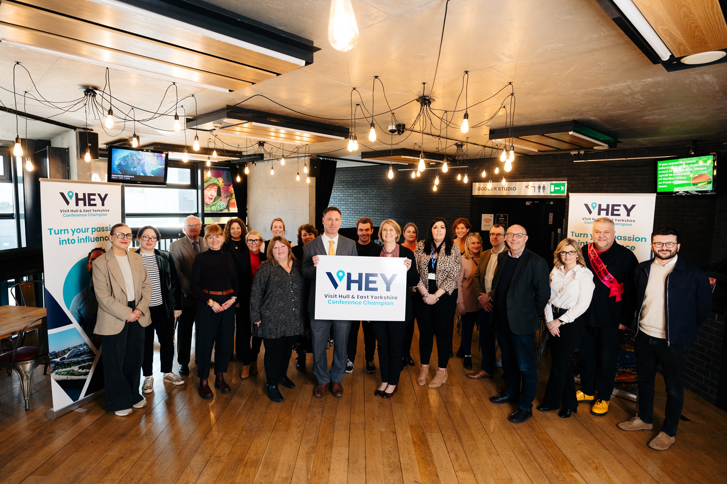 Conference Champions gathered in Hull Truck Theatre to celebrate launch. There is a mix of ages and genders. Everyone is smiling and in the centre a delegate is holding a white board with "VHEY Conference Champions" branded in blue.